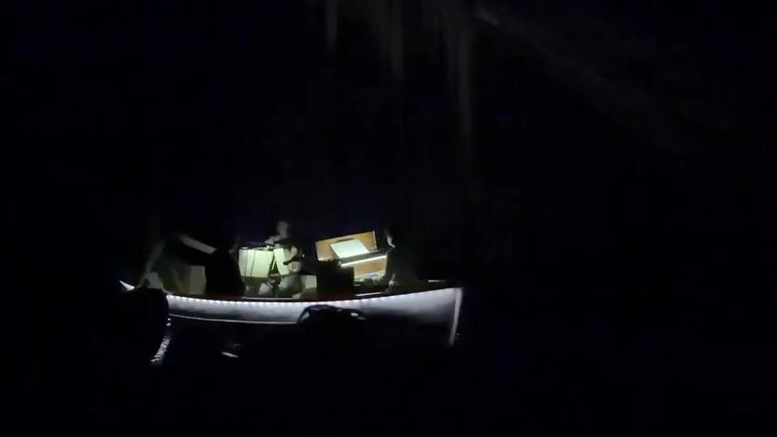 A low-lit, dark scene shows a small boat with at least three people inside, moving slowly on water in a cave. The boat has LED strip lights along the rim, providing the main source of illumination. There's some equipment in the boat, including what appears to be a laptop or screen in front of one of the passengers. The background is largely darkness, but there are some indistinct cave formations visible above the boat. One person is actively rowing. The atmosphere seems quiet and exploratory.