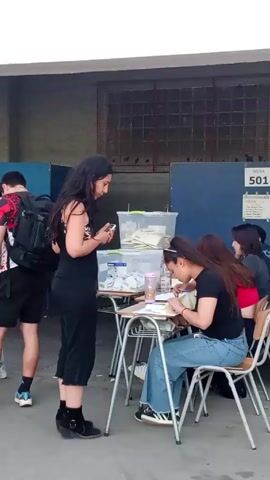 A woman in a black tank top and cropped pants looks at her smartphone while standing near a table where another woman, wearing a black top and jeans, sits and writes on a paper. There are several plastic containers with papers on the table, and a sign with "Mesa 501" is visible behind the table. Other individuals are seated or walking nearby, and the setting appears to be an outdoor area possibly related to voting or registration.