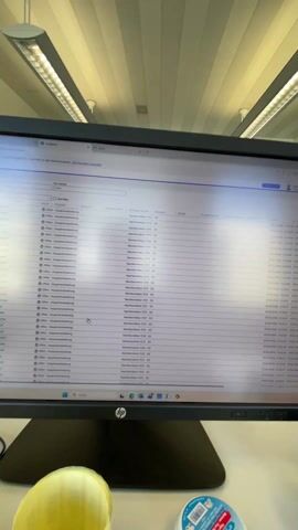 The video shows a computer monitor displaying a grid of data. The monitor is on a desk with office supplies. The camera then pans back to reveal a laptop and headphones on a white desk. Finally, the camera focuses on a man wearing a white shirt with black abstract patterns. He is looking into the camera with his eyes closed, seemingly disinterested or bored. Behind him is a window.