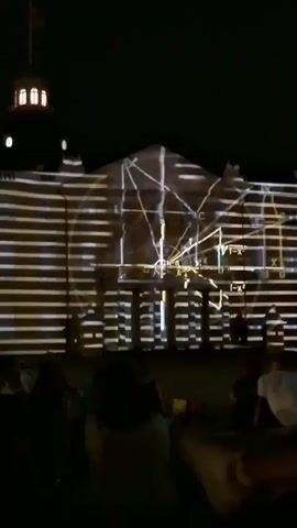 The video captures a nighttime outdoor projection mapping event. A large building facade is illuminated with abstract, dynamic light patterns. The projections consist of horizontal lines that shift and undulate, creating a sense of movement and depth. In some segments, geometric shapes and diagrams, possibly related to mathematics or science, are also projected onto the building. The scene is viewed from a distance, with the silhouettes of an audience sitting or lying on the ground in the foreground, suggesting a public gathering or performance. The overall atmosphere is dark, with the building and its light show being the primary focal point.