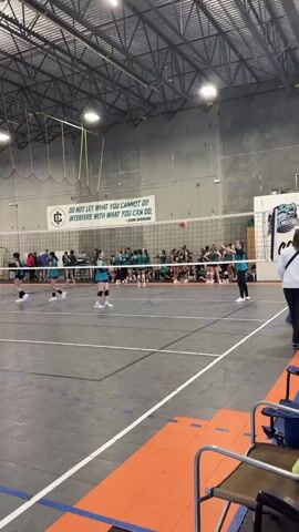 The video shows a group of young women playing volleyball in a large indoor sports facility. They are wearing matching teal uniforms. The facility has a high ceiling with exposed metal beams and multiple overhead lights. There are spectators sitting on benches along the sidelines. Banners with sports team logos hang on the walls. The volleyball court has clearly marked lines. The camera angle offers a full view of the court and the players' movements during the game.
