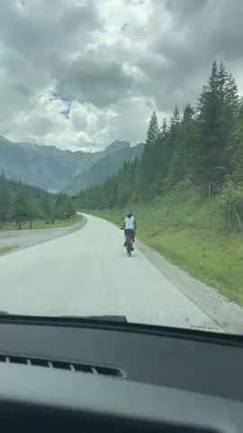 A video taken from inside a car shows two people on bicycles riding on a paved road through a mountainous, forested area. The road curves to the left. The sky is cloudy. On the right side of the road, there is a fence and trees. On the left side, there are trees and mountains in the distance. The car is moving forward.