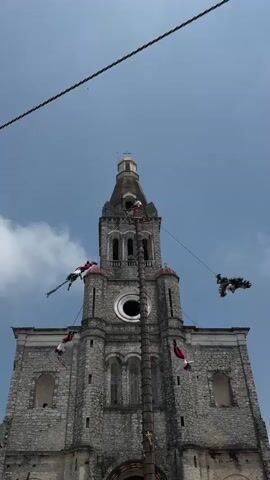 The scene is a clear day with a large stone church in the background. In front of the church is a tall, wooden pole. Four people in red and white costumes are suspended from the pole by ropes. They are twirling or spinning in the air. One of the figures appears to have wings. There is a thin wire running across the top of the frame.