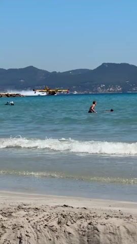 The video captures a bright, sunny day at a beach in La Ciotat, France. A large, yellow twin-engine seaplane, characteristic of a firefighting aircraft, is seen skimming across the surface of the turquoise water from left to right, creating a significant white spray wake behind it. The plane appears to be either practicing a water scoop or taking off after collecting water.

In the mid-ground, several individuals are in the ocean. A man in dark swim shorts stands waist-deep in the foreground, facing right and watching the plane. Another person is visible further out, swimming. A third, more distant swimmer, is also present. The water is a clear, inviting blue with gentle waves breaking on the sandy shore in the immediate foreground.

Beyond the water, in the distance, a range of green, tree-covered mountains stretches across the horizon under a clear, bright blue sky. Scattered buildings are visible on the slopes of these hills. The overall impression is one of a busy, yet picturesque summer day. The audio includes the distinct roar of the seaplane's engines and excited exclamations, possibly from children, indicating their fascination with the passing aircraft. There is no visible text.