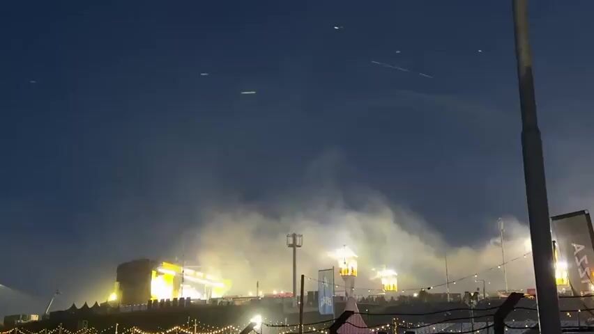 A static, wide outdoor shot at a festival shows a stage on the left, illuminated by changing red and white lights and accompanied by smoke. Two tall light towers are visible, one in the middle and another on the right, also emitting light and smoke. The sky is dark, and string lights are strung across the bottom of the frame. In the distance, streaks of light appear to be flying across the dark sky.