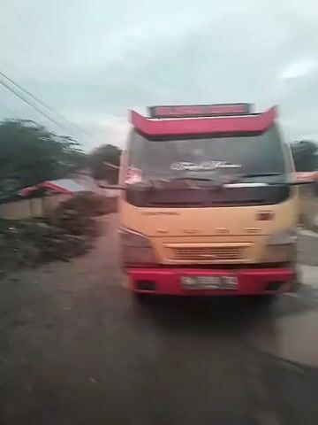 The video shows a yellow and red Mitsubishi Colt truck driving on a wet, muddy road. The truck has a red sun visor with white lettering "EURO DAI" on top. A sticker on the windshield reads "Tiga Manis". The truck's license plate is visible: "BM 8398 TQ". The road is lined with vegetation and a few simple structures, possibly shacks. Power lines are visible in the overcast sky. The camera is positioned in front of the truck, moving slightly as it records.