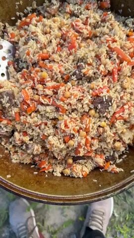 This video shows a close-up, overhead view of a large metal pot filled with pilaf. The pilaf appears to be cooked with rice, chunks of dark meat, julienned carrots, and chickpeas. The pot is a rustic, dark-colored cauldron-like vessel. The food is being stirred or scooped with a utensil visible in the upper left corner and in the final frame where a forkful of pilaf is lifted. In the lower portion of the frame, the legs and feet of a person wearing casual clothing, including light-colored athletic shoes, are visible, indicating they are standing and holding the camera or observing the food. The background is slightly blurred and appears to be outdoors with some green foliage suggesting grass or trees. The lighting suggests natural daylight.