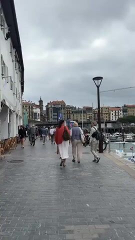 The video shows a paved walkway alongside a body of water, likely a harbor or marina, with buildings in the background. Several people are walking along the path. In the foreground, three women are walking away from the camera. The woman in the center has blonde hair and is wearing a light-colored outfit. The woman to her left is wearing a red jacket and a white skirt. The woman to her right has blonde hair and is wearing a light-colored outfit and a backpack. There is a lamppost on the right side of the walkway, and a bicycle is parked next to it. In the background, there are boats docked in the water and a crane visible. The sky is cloudy.