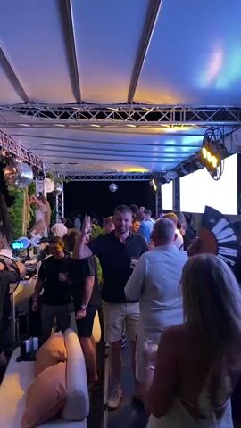 This media file depicts a vibrant outdoor party scene at night, likely in Baan Chaweng, Thailand. The setting is an open-air venue with a white canopy overhead, illuminated by warm yellow and orange lights, creating a festive atmosphere. Several disco balls hang from the structure, reflecting the lights and adding to the celebratory mood. Lush green foliage is incorporated into the decor, trailing from the ceiling, and white spherical decorations are also visible.

The foreground features a lounge area with a white couch adorned with large, plush cushions in shades of yellow and beige. In the midground and background, a crowd of people is actively engaged in a party. Many individuals are dancing, with some raising their hands in the air. The overall mood appears to be energetic and joyful.

Notable individuals include a man in the center wearing a black polo shirt and beige shorts, actively interacting with the crowd, and a woman in a long, flowing white dress on the left. Other attendees are dressed in casual attire suitable for a warm evening.

The time of day is clearly night, as indicated by the artificial lighting and the dark sky visible beyond the canopy. There are no specific weather cues, but the outdoor setting suggests a pleasant evening.

Visible text includes "RIVA STARR" displayed prominently on a screen or backdrop, suggesting a DJ or performer associated with this name might be present or featured. The presence of multiple disco balls, the music, and the dancing crowd all point to a lively event, possibly a club night, private party, or festival.