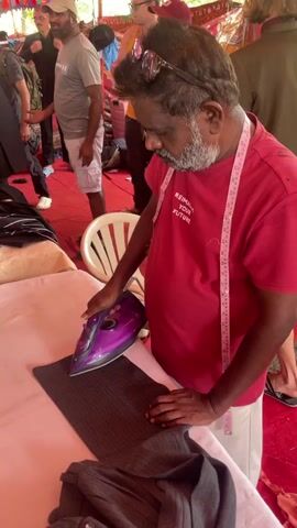 The video shows a man ironing a pair of dark gray or black pants on a white surface. The man is wearing a red t-shirt with the words "REIMAGINE YOUR FUTURE" printed on it, white shorts, and a measuring tape around his neck. He has sunglasses on his head. He is using a purple and black iron. Several people are visible in the background, some standing and some moving. A red tarp or rug covers the ground. A white plastic chair is near the ironing surface. There are various articles of clothing near the man and on the ironing surface. The environment appears to be under a tent or temporary shelter with decorative elements.