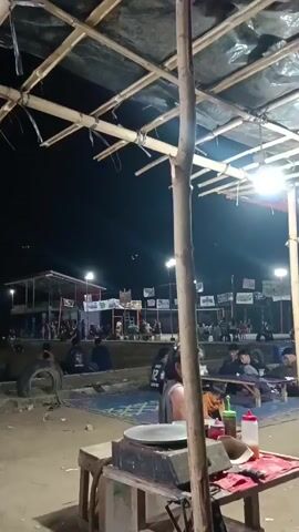 A nighttime outdoor scene depicts a gathering of people, possibly at an event or market, illuminated by artificial lights. In the foreground, a wooden table with a cooking apparatus, possibly a griddle, is visible. Behind it, a thin wooden pole supports a makeshift roof constructed from bamboo and dark plastic sheeting. A group of people are seated or standing further back, some facing a lit area where other individuals appear to be engaged in some activity. Banners and flags are strung across poles in the middle ground. The overall environment appears to be somewhat rustic, with dirt or sand on the ground.