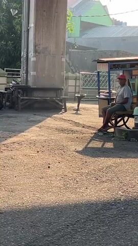 The video shows a man sitting on a stool under a raised structure, possibly a bridge or an overpass, in what appears to be an outdoor seating area. He is wearing a red cap, a grey t-shirt, and dark shorts. The ground is covered in dirt and gravel, and there are shadows cast by the structure. In the background, there is a small food stall or cart with a blue awning. The overall environment suggests a street or an informal commercial area. The man is looking towards the left side of the frame and appears to be relaxed.