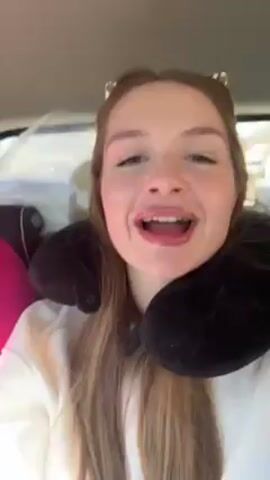 A young woman wearing a black neck pillow and a white sweater is singing in the car. She smiles and moves her hands while singing. She has red nail polish. The car seems to be moving, and there are bags and luggage in the back.