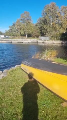 The video shows a sunny day at a lake. The camera moves from an upside-down yellow canoe on a grassy bank, past rocky shoreline, over a concrete block, and along a wooden dock with a white metal ladder, finally focusing on the rippling blue water. Trees line the opposite shore, and several buildings are visible. Boats are docked in the distance. The sky is clear blue, with a few thin clouds appearing later. The only person visible is a shadow cast by the camera operator near the canoe.