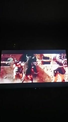The video shows a scene from a movie or TV show. Several people dressed in ancient Roman-style armor are fighting in what appears to be an arena or combat setting. The scene is fast-paced and chaotic, with dust and debris kicked up from the ground. One person is wielding a large axe, while others are using swords and shields. There's a lot of movement and action, suggesting a battle is taking place. The lighting is dramatic, with bright highlights and deep shadows.