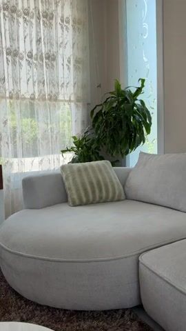 The media file shows a living room scene with a focus on a plush, cream-colored sectional sofa. A textured throw pillow with green stripes rests on the sofa. Behind the sofa, sheer curtains with a floral pattern hang over a window, revealing a glimpse of greenery outside, suggesting a daytime setting. A large houseplant is placed to the right of the window. To the left of the window, a white cabinet with a wooden front, decorated with a geometric pattern, is visible. On top of the cabinet, a gold vase holds dried flowers, and there are also decorative candle holders and a small green dish. A tall, thin object with a blue cap, possibly a bottle, is in the foreground, slightly out of focus. A portion of a television screen displaying a colorful image is visible on the left side of the frame. The overall ambiance is calm and well-lit, indicating a comfortable interior space. There is no visible text in the media file.
