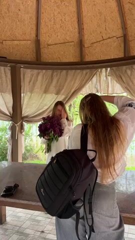 Inside a circular wooden gazebo, two women are present. One woman, dressed in a white blouse, stands facing the other, who is presumed to be a photographer. The woman in white holds a large bouquet of purple lilacs and covers her right eye with her hand. She is smiling. The presumed photographer is also wearing a white blouse and light-colored jeans. She has long, light brown hair and is holding a black backpack. On a dark table in front of them sits a pair of sunglasses. Outside the gazebo, foliage and other vegetation are visible.