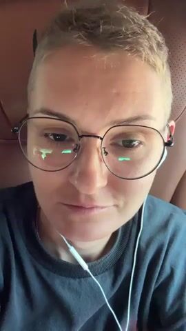 A close-up selfie video shows a person wearing round glasses and white earbuds, looking directly at the camera. The person is seated in what appears to be a bus or van, with brown leather seats visible in the background. The camera pans slightly to reveal other passengers in the vehicle, all looking at their phones. One person in the background is wearing a white shirt, another a black shirt, and a third a red shirt. The lighting suggests it is daytime, possibly inside a vehicle with windows reflecting the outdoors. The video is short and static, with no discernible actions or interactions between the individuals.