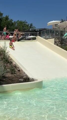 A boy in a yellow swimsuit slides down a water slide and splashes into a pool. He is followed by another boy in a black swimsuit. In the background, other people are seen near the top of the water slide, and some are in the pool. The scene takes place on a sunny day, with clear blue skies and lush green foliage surrounding the water park.