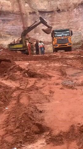 An excavator is seen digging in a quarry with a large dump truck waiting nearby. Three individuals are standing between the excavator and the truck. The ground is covered in red dirt, with tire tracks visible in places. The excavator's arm is extended and its bucket is full of dirt. The truck's headlights are on. One of the individuals is wearing a reflective vest.