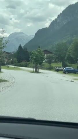 This is a video recorded from inside a vehicle looking out at a mountainous landscape in Flachauwinkl, Austria. The scene shows a paved road curving to the left, bordered by a cobblestone area with several parked bicycles. A large white truck with text on its side is visible on the left.

In the background, there are buildings, scattered trees, and imposing green mountains under a cloudy sky. A few people are seen near the bicycles and the truck. The weather appears overcast, suggesting it might be morning or afternoon, with no direct sunlight. The overall atmosphere is peaceful and rural, with the natural beauty of the mountains dominating the scene.

The text on the white truck reads "Weiss & Nesch" followed by "www.weiss-nesch.at". The video captures a serene moment in a mountainous region, likely a stop for travelers or a local gathering point for cyclists.