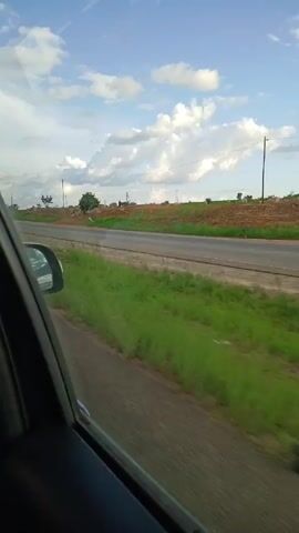 The view from a car window shows a highway flanked by green grass. A shallow, elevated embankment runs parallel to the road, topped with sparse vegetation and a few small trees.  The sky above is blue with scattered white clouds.  A truck passes in the opposite direction.