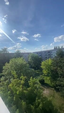 The video pans across a residential area with apartment buildings and trees under a bright, partly cloudy sky. In the foreground, a white balcony railing is visible. Beyond the trees, a long, light-colored apartment building with multiple balconies can be seen. A path winds through a grassy area with more trees, including some coniferous trees. A person is walking on the path in the distance. The sun is visible in the upper left corner, casting lens flare.