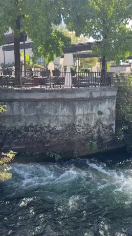 The video shows a close-up view of a rushing river with choppy water. Above the river, a stone wall with a weathered appearance is visible. Behind the wall, a patio area of a building is seen with tables, chairs, and large, white, floor-standing lamps. Several trees with green leaves are situated in front of the patio, casting shadows. The sky above is bright and appears to have some scattered clouds. The scene conveys a natural outdoor setting combined with an outdoor dining or seating area.