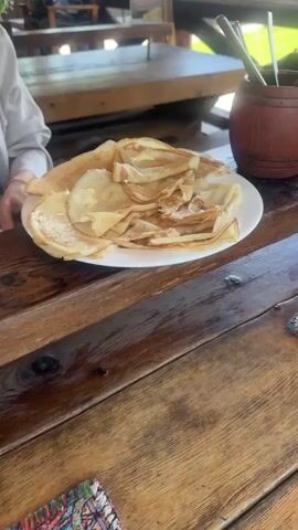 The video shows a plate of pancakes on a wooden table. Some of the pancakes are folded into triangles. There are small dots of butter melting on the pancakes. A person wearing a light-colored shirt is partially visible in the background to the left of the plate. Behind the plate, there is a wooden container filled with utensils and another wooden table. The overall lighting is slightly warm and natural.