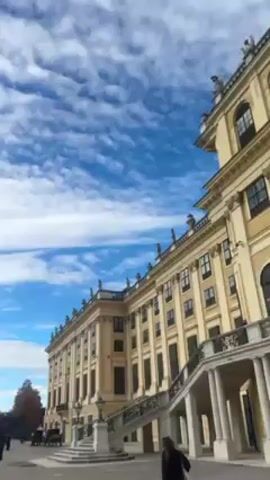 The video shows a low angle view of Schönbrunn Palace in Vienna, Austria. The sky is blue with white clouds. People are walking around the palace grounds. The video pans around showing the large open area in front of the palace with a gravel path and trees on the side. As the camera pans around, the sky becomes more cloudy.