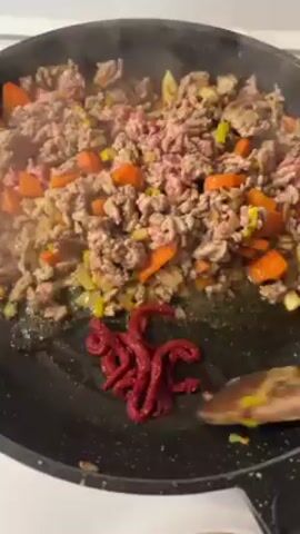 In a close-up shot, a black frying pan sits on a white stovetop, filled with steaming ground meat, diced carrots, onions, and leeks. A wooden spoon is used to stir in a dollop of dark red beet paste.