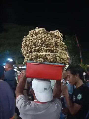 A man wearing a white cap carries a large basket of peanuts on his head, walking through a crowded outdoor market at night. The basket is red and overflowing with peanuts. Several people surround him, some looking at the peanuts, others talking. One man in the foreground is drinking from a bottle. The background is dark with some trees and lights visible. There are flags and stalls suggesting a festive or market atmosphere.