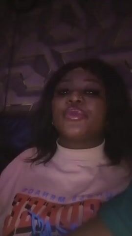 The video shows two people in what appears to be a dark, possibly dimly lit indoor space. The first person, a woman with dark hair, is wearing a light-colored turtleneck shirt with a graphic on it. She has visible makeup, including glossy lips, and a nose ring. The second person is a man wearing a green shirt. The environment has a textured or patterned wall in the background, potentially suggesting a bar or club setting, based on the music. The video appears to be taken from a close perspective, focusing on the individuals, and the man gives a brief smile to the camera.