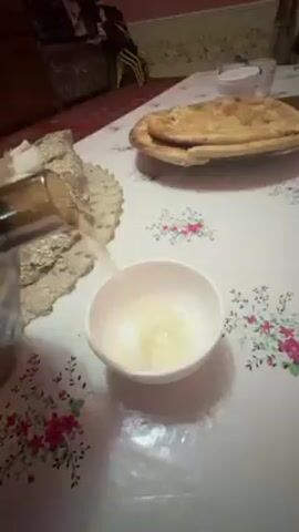 A hand pours a clear, yellowish liquid from a gold-colored teapot with a faceted design and a crystal knob on the lid into a small white bowl on a white floral tablecloth. Steam rises from the spout of the teapot. A plate of flatbread sits on the table. The background shows a room with furniture and a red patterned rug. The voice of a child can be heard.