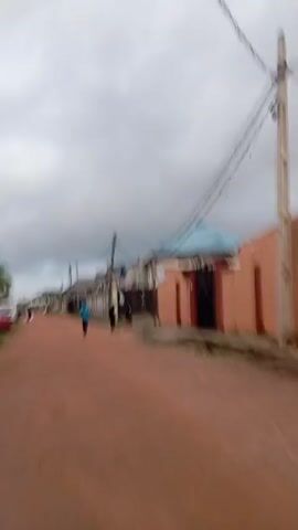 This media file depicts a street scene in Idimu, Nigeria. The road is unpaved and made of reddish-brown dirt. On either side of the street, there are buildings with various colored walls, including orange, red, and white. Some buildings have blue roofs. Overhead, there are utility poles with numerous black wires crisscrossing the sky.

Several people are visible walking along the street. In the foreground, a person in a blue top and dark pants walks away from the camera. Further down the street, other figures can be seen, including someone on a bicycle and others walking. There are also a few cars parked along the side of the road, including a red car and a white van.

The sky is overcast with gray clouds, suggesting it might be a cloudy day or possibly early morning or late afternoon. The overall atmosphere appears to be that of a typical residential street in a community. No specific event or activity is clearly identifiable beyond people moving about their daily routines. There is no visible text in the media file.