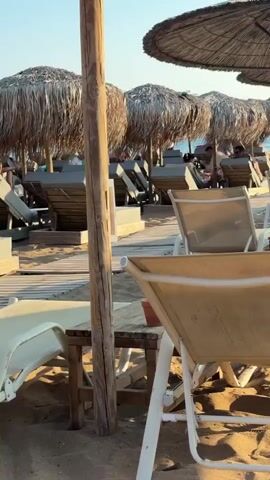 The video pans across a sandy beach with rows of empty lounge chairs and thatched umbrellas. Wooden walkways are laid out on the sand. In the distance, a person sits in a lounge chair under a colorful umbrella. The sea is visible beyond the beach, and the sky is clear and bright, suggesting a sunny day. The sun casts shadows across the sand and chairs.