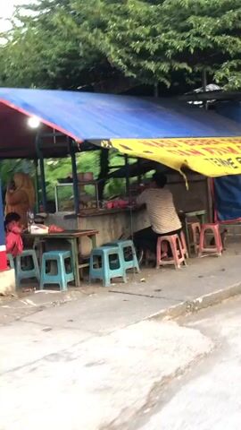 The video shows a street food stall with a blue tarp covering a yellow awning. Several blue plastic stools and two pink stools are arranged in front of the stall. A person wearing a striped shirt is seated on one of the pink stools, facing away from the camera and appearing to be working at the stall. Various items are visible on shelves behind the stall, including what looks like food in containers and bags. The ground in front of the stall is paved. The overall impression is of a modest, outdoor food vendor.