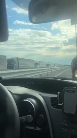 The camera films from inside a car driving on a highway. On the other side of a concrete barrier is a line of semi-trucks. The sky is blue with white clouds. The car's speedometer reads 134 kilometers per hour. A phone mount is visible on the dash. Music is playing.