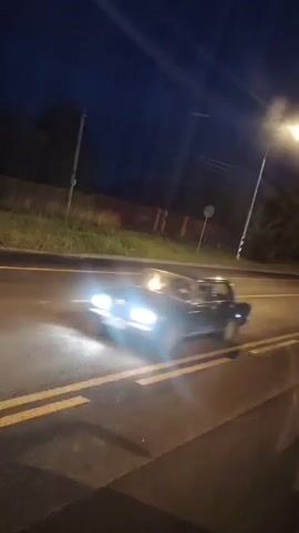 The video captures a nighttime scene on a road. A streetlamp illuminates the road and surrounding grassy area. There are double yellow lines painted on the asphalt. A car with its headlights on drives by, creating a blur of motion. The sky is dark blue, indicating it is either dusk or nighttime. A tall pole, possibly a utility pole, is visible on the left side of the road. A red fence and some trees are visible in the background.