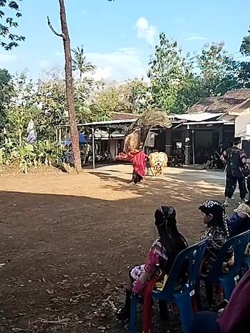 The video shows a dancer in a traditional costume performing in an outdoor area. The dancer is wearing a red and black outfit with a large, ornate headpiece featuring tall, dark feathers. The dance is taking place on a dirt surface, and several people are watching from blue plastic chairs in the foreground. Behind the dancer is a covered structure with people seated inside. There is a sound system and what appears to be a mask on a stand nearby. The environment includes trees, plants, and buildings in the background, with a partly cloudy sky above. The video is taken from a stationary position, showing the dance performance in a wide shot.