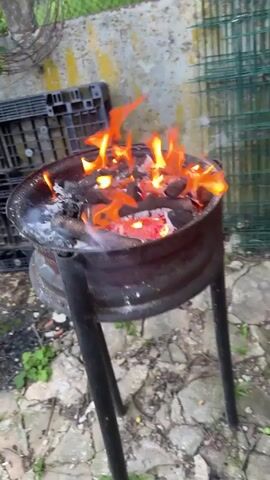 The video shows a close-up shot of a makeshift barbecue grill. The grill appears to be constructed from a metal rim mounted on black metal legs. Inside the rim, burning charcoal and wood logs are visible, with flames licking upwards. Embers glow red beneath the charcoal. In the background, out of focus, are various objects including what appears to be a black plastic crate, a weathered wall, and some green fencing. The ground beneath the grill is covered in rough stones or gravel. The lighting suggests an outdoor daytime setting. There are no people or animals visible in the frame.
