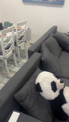 The camera pans around a sparsely decorated room, starting on a gray sectional sofa with a large panda plush toy resting on it. The room has light-colored wood flooring. The camera then moves to show a white wooden chair with a laptop sitting on it. A television is mounted on the wall above the chair. The television screen displays a YouTube video. A pair of slippers sit on the floor near the chair. There are two dark-colored doors on the left side of the frame. The ceiling appears to have a design resembling clouds. The room looks like a casual living or entertainment space.