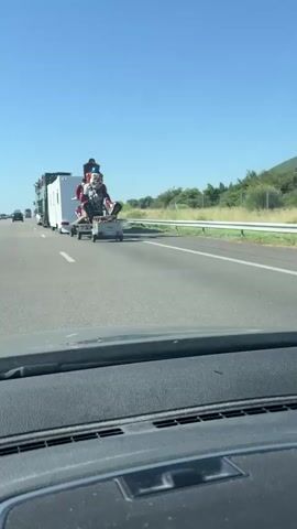 A tall, elaborate clown figure is being transported on a trailer on a highway. The clown is brightly colored with red and blue accents and appears to be sitting down. It is attached to a white trailer, which is then hitched to a larger vehicle, possibly a truck, further up the road. The trailer also carries a smaller, light-colored utility vehicle. The scene takes place on a sunny day with a clear blue sky and a few green trees visible on the side of the road. The perspective is from inside another vehicle, looking out at the clown and trailer on the highway.