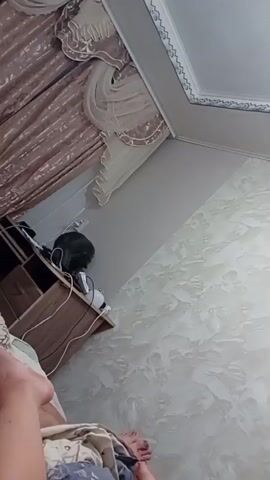 The video shows a domestic interior scene, possibly a bedroom. The camera is positioned at a low angle, looking towards a wall with textured wallpaper. In the background, a curtain is visible, drawn to the left. A shelf unit is positioned against the wall, and a cat is sitting on top of it, looking downwards and to the right. The cat is grey and appears to be grooming itself. There are some objects on the shelf, including what looks like an electronic device. In the foreground, a person's legs and feet are visible, covered partially by bedding. The lighting suggests it is daytime, and the overall impression is one of a casual, everyday moment.