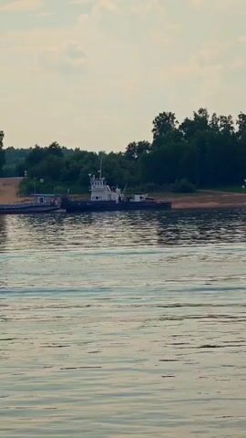 This is a video clip showing a ferryboat on a wide river. The ferry is a utilitarian vessel, dark in color with a white superstructure, carrying a few vehicles, including cars and possibly a truck. It appears to be docked or about to dock at a ramp on the shore. The shore is lined with lush green trees, and beyond that, there is a light blue sky with scattered clouds, suggesting it is daytime. The water of the river is calm, with gentle ripples reflecting the light. The overall scene is peaceful and suggests a quiet, possibly rural setting in Erkalovo, Russia, where ferry crossings are a common mode of transportation. The time of day appears to be late afternoon or early evening due to the soft lighting and the color of the sky.