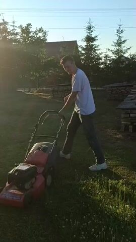 This media file captures a young man mowing a lawn in a rural setting. He is dressed in a white t-shirt and dark pants, pushing a red lawnmower. The sun is setting, casting long shadows and bathing the scene in a warm, golden light, suggesting it is late afternoon or early evening. The weather appears to be clear and pleasant.

The background reveals a residential property with a house visible through trees. There are also stacks of lumber or wood to the right, hinting at ongoing construction or DIY projects. The lawn is green and appears to be well-maintained, though there are patches of taller grass and weeds. The trees are predominantly coniferous, and the overall atmosphere is peaceful and serene.

The man's posture and the way he is operating the lawnmower suggest he is engaged in a routine chore. There are no obvious signs of distress or excitement; his expression is neutral as he focuses on the task. The scene evokes a sense of domesticity and the simple pleasures of maintaining one's home and surroundings in a tranquil, rural environment. The location context of Zakrzewo-Kopijki, Poland, further grounds the scene in a specific Eastern European rural landscape.

There is text visible on the lawnmower, but it is too blurry to be read clearly. However, the red color and branding suggest it's a common consumer model. The overall impression is one of a quiet, ordinary moment in a countryside setting.