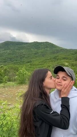 The video presents a view of a mountainous landscape on a cloudy day. Initially, two people, a woman with long brown hair and another person partially visible, are seen in the foreground, appearing to share a kiss. The camera then pans upwards to reveal a lush, green mountain covered in trees under an overcast sky filled with dark, heavy clouds. Small flying insects are visible against the sky. The landscape appears peaceful and natural, despite the looming weather.