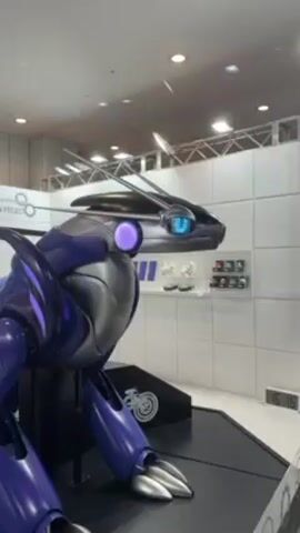 A robot, designed to resemble the Pokémon Miraidon, is on display at an event. The robot is primarily purple and gray with a sleek, futuristic design. It has a central purple orb and blue, glowing eyes. The robot is presented on a black platform, situated in front of a white wall. A Pokémon logo and the words "TOYOTA ENGINEERING MIRADON PROJECT" are on the wall behind the robot. The setting appears to be an indoor exhibition or convention space.