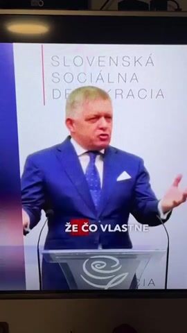 A man in a blue suit and tie stands behind a podium with a microphone. He is speaking and gesturing with his hands. The podium has a logo on it. Behind him, a screen displays text that reads "SLOVENSKÁ SOCIÁLNA DEMOKRACIA". There are also two lines of text overlaid on the video in white and red, reading "ŽE ČO VLASTNE" and "ALEBO NEPRIJALO".