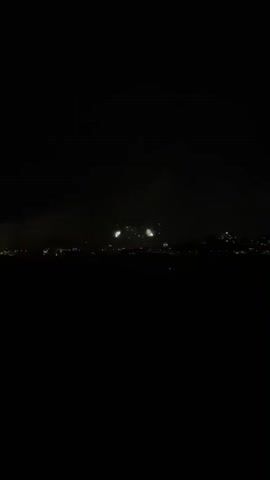 A night time video captures a distant cityscape with fireworks. The sky is dark with a band of clouds faintly visible. Two clusters of fireworks explode, one in white and green, the other in pink and yellow. The city lights are a blurry orange glow beneath the fireworks. The video has a slightly shaky quality.